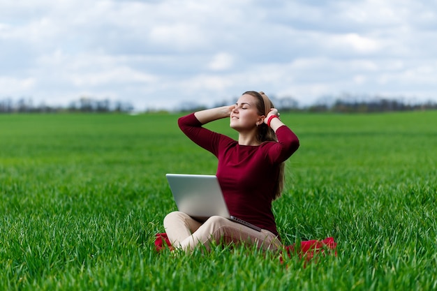 Mujer de éxito joven está sentada sobre la hierba verde con una computadora portátil en sus manos. Descanse después de una buena jornada laboral. Trabaja en la naturaleza. Chica estudiante trabajando en un lugar apartado. Nuevas ideas comerciales