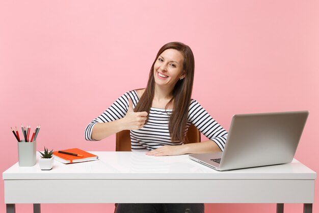 Mujer de éxito joven que muestra el pulgar hacia arriba sentarse y trabajar en el escritorio blanco con un ordenador portátil contemporáneo aislado sobre fondo rosa pastel. Concepto de carrera empresarial de logro. Copie el espacio para publicidad.