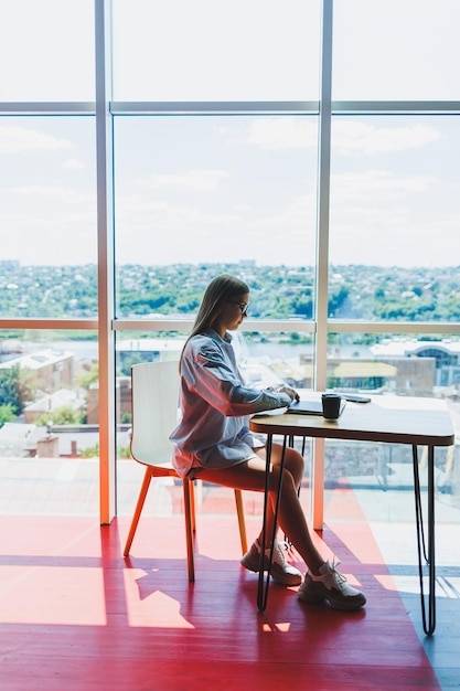 Mujer exitosa está mirando una computadora portátil en un café y tomando café Una mujer sonriente joven con gafas se sienta en una mesa cerca de la ventana con un teléfono Trabajo independiente y remoto Estilo de vida femenino moderno