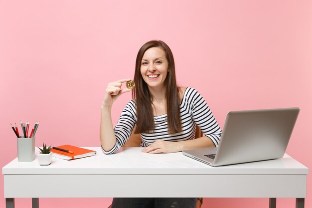 Mujer exitosa sosteniendo bitcoin, moneda de metal de color dorado, futura moneda sentarse a trabajar en un escritorio blanco con computadora portátil pc aislada sobre fondo rosa pastel. Concepto de carrera empresarial de logro. Copie el espacio.