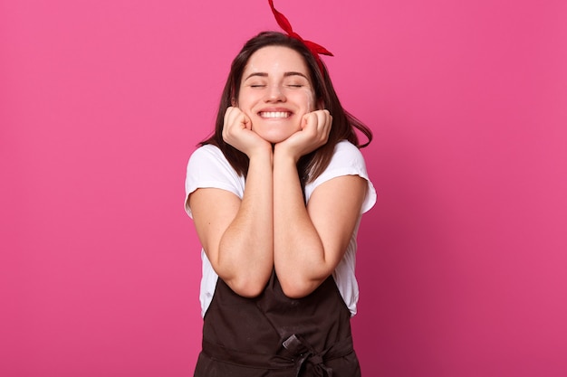 Mujer con expresión facial agradable, poniendo las manos en la cara, cerrando los ojos, sonriendo sinceramente