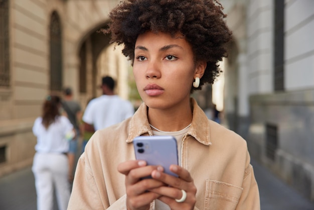 mujer con expresión seria usa un teléfono inteligente para las redes sociales páginas de redes sociales paseos al aire libre explora la ciudad usa una chaqueta beige informal comparte multimedia en un blog