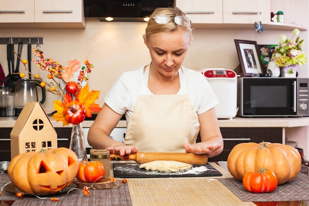 Una mujer extiende masa para galletas para Halloween con un rodillo en la cocina con una decoración otoñal. Haciendo galletas para Halloween.