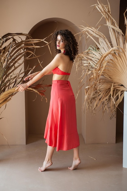 Una mujer con falda roja se encuentra en una habitación con una palmera al fondo.