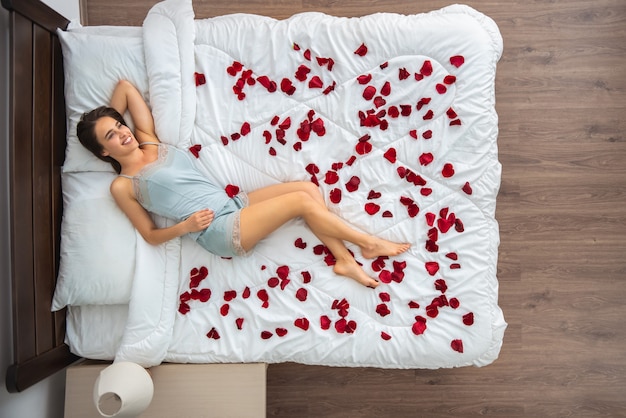 La mujer feliz acostada en la cama con pétalos de rosa. vista desde arriba