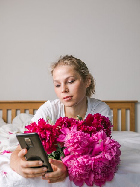 Mujer feliz acostada en la cama en pijama, disfrutando de un ramo de flores de tulipán de peonía y usando el teléfono