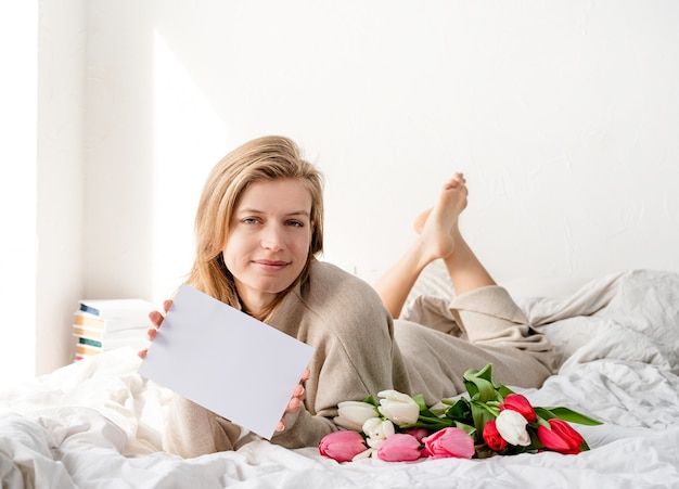 Mujer feliz acostada en la cama en pijama, sosteniendo un ramo de flores de tulipán y una tarjeta en blanco para un diseño simulado