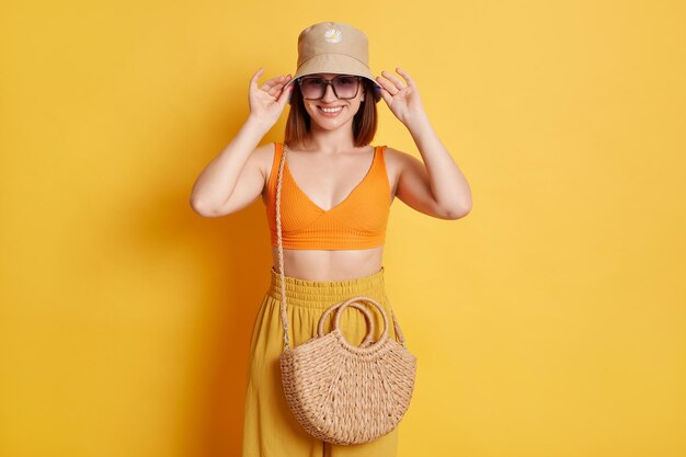 Una mujer feliz y alegre con una camiseta naranja y gafas de sol aisladas sobre un fondo amarillo tomándose de la mano el sombrero mirando sonriendo a la cámara