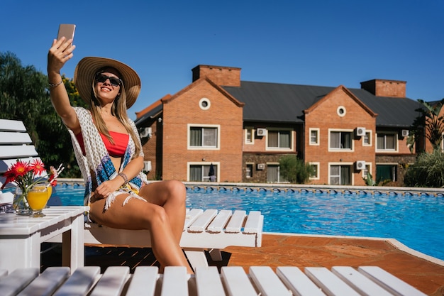 Foto mujer feliz y alegre sentada en una tumbona cerca de la piscina y tomando un selfie en su teléfono celular.