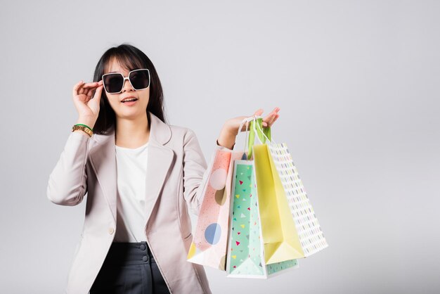 Mujer feliz con anteojos comprador seguro sonriendo sosteniendo bolsas de compras en línea colorido multicolor, retrato emocionado asiático joven mujer compra estudio tiro aislado sobre fondo blanco, venta de moda