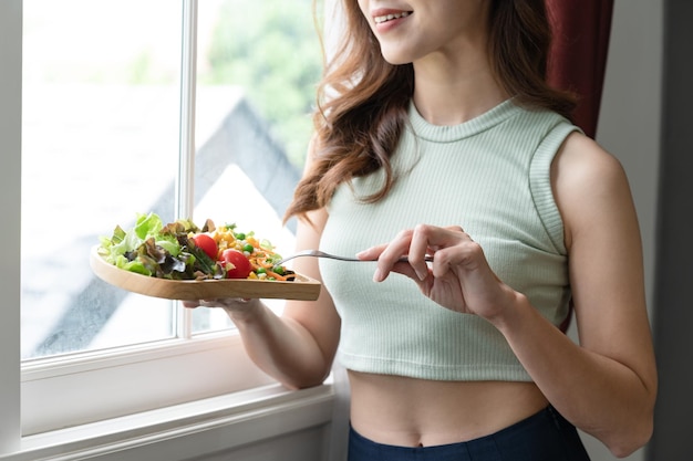 Mujer feliz de Asia comiendo ensalada con luz solar de ventana en la sala de estar