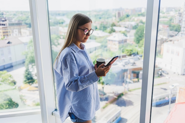 Mujer feliz bebe café y mira por la ventana en un café Una joven sonriente con gafas está de pie junto a la ventana Ocio recreativo y tiempo libre El estilo de vida independiente moderno