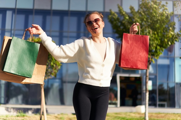 Mujer feliz con bolsas de compras caminando en la ciudad desde el centro comercial Chica emocionada disfruta de las compras en la venta del viernes negro Mujer posando en cámara con bolsas de papel llenas Concepto de consumismo