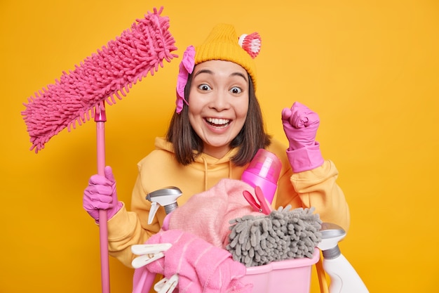 Mujer feliz con cabello oscuro aprieta los puños se siente feliz de usar detergentes efectivos y detergente en polvo para limpiar el polvo en la habitación haciendo lavandería sostiene fregona aislada sobre pared amarilla