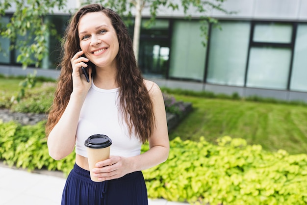 Mujer feliz con café para hablar por teléfono a pie en la calle de la ciudad