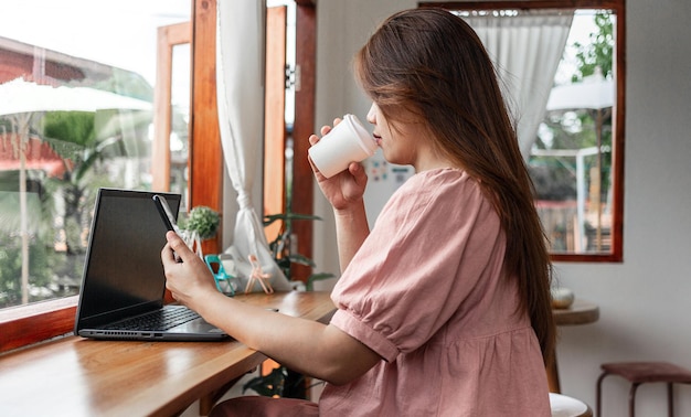 Una mujer feliz en un café usando un teléfono inteligente y una computadora portátil tomando café en una taza de papel en el café