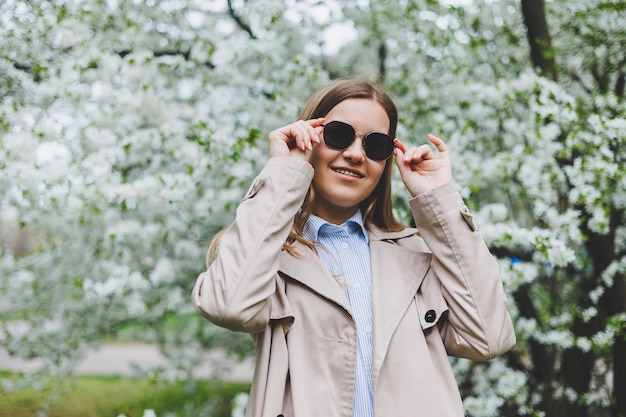 Mujer feliz caminando usando un teléfono inteligente en una calle de la ciudad Mujer de negocios con gafas de sol usa un teléfono móvil al aire libre bajo un árbol floreciente chica escribiendo un mensaje charlando en la página web mirando a la cámara