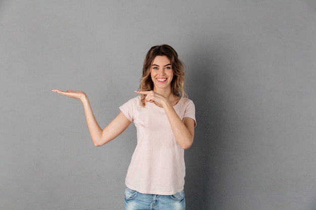 Mujer feliz en camiseta con copyspace en la palma y apuntando sobre ella mientras sobre gris