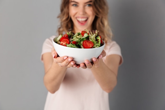 Mujer feliz en la camiseta que muestra la placa con las verduras frescas sobre gris. Centrarse en la placa