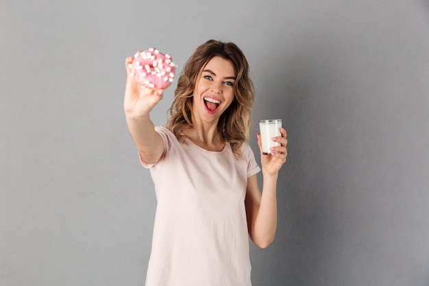 Mujer feliz en la camiseta que sostiene la taza de leche mientras que muestra el buñuelo. Centrarse en donat