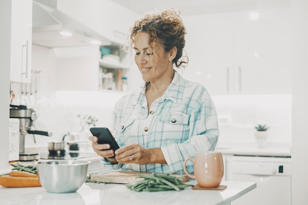 Mujer feliz en casa preparando el almuerzo y mirando el teléfono móvil para seguir instrucciones o tutoriales o recetas de nuevas ideas Mujeres modernas en línea charlando solas Usando celular en el ocio interior