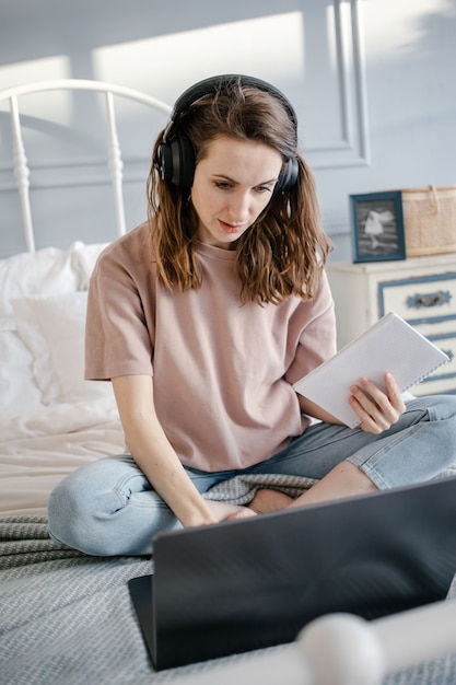 Mujer feliz en casual con los auriculares trabajando en una computadora portátil de forma remota desde casa en la cama