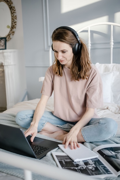 Mujer feliz en casual con los auriculares trabajando en una computadora portátil de forma remota desde casa en la cama