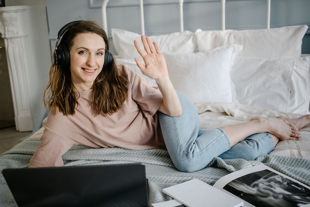 Mujer feliz en casual con los auriculares trabajando en una computadora portátil de forma remota desde casa en la cama