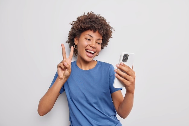 Una mujer feliz y complacida se toma una selfie o hace gestos de videollamada, el signo de la paz sostiene el teléfono móvil y usa una camiseta azul casual aislada sobre fondo blanco. Concepto de lenguaje corporal de tecnología de personas