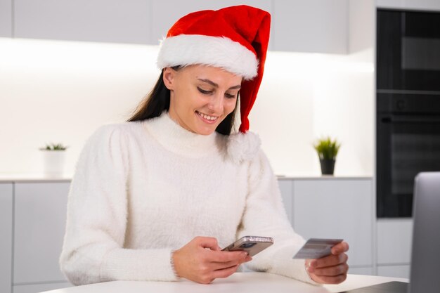 Mujer feliz comprando regalos en línea sentada en la cocina moderna mujer en sombrero de Santa Claus gastando