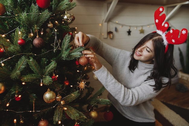 Mujer feliz en cuernos de reno decorando el árbol de navidad con adorno dorado en la sala festiva