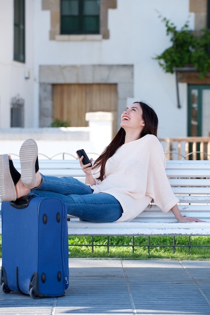 Mujer feliz de cuerpo completo con maleta y celular en banco de parque