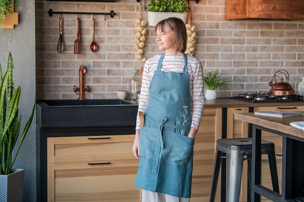 Mujer feliz en un delantal de lino de pie en la cocina en la mesa con una sonrisa mira por la ventana