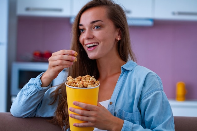 Mujer feliz descansando, riendo y comiendo crujientes palomitas de caramelo durante ver películas de comedia en casa. Película de palomitas de maíz