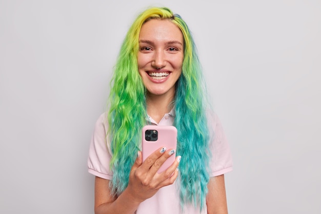 Mujer feliz de descargar la aplicación en el teléfono inteligente sonríe felizmente tiene el pelo largo teñido piercing en la nariz vestidos casualmente aislado en gris