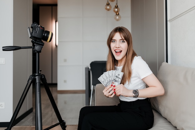 Mujer feliz con dinero grabando video tutoriales para blog en línea en Internet en casa en el sofá