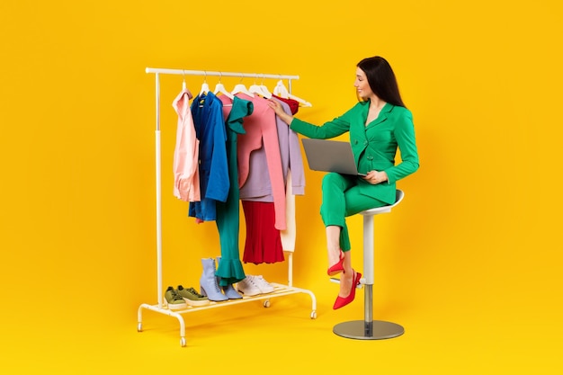 Mujer feliz diseñadora de moda con una computadora portátil mirando ropa en un estante de warbrobe usando una computadora portátil