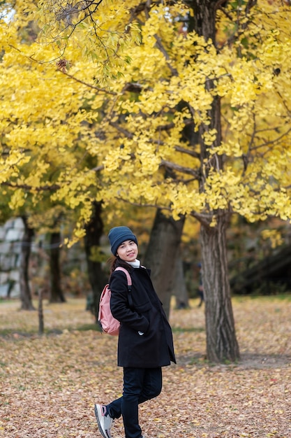 Mujer feliz disfrutar en el parque al aire libre en la temporada de otoño, viajero asiático en abrigo y sombrero contra el fondo de hojas amarillas de Ginkgo