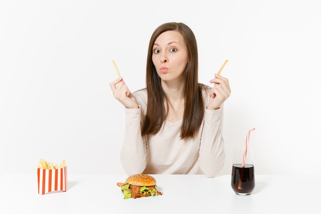 Mujer feliz divertida en la mesa con varios trozos de patatas, papas fritas, hamburguesas, cola en botella de vidrio aislado sobre fondo blanco. Nutrición adecuada o comida rápida clásica estadounidense. Área con espacio para copiar.