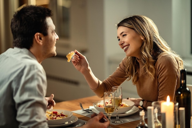 Mujer feliz divirtiéndose mientras alimenta a su novio durante la cena en la mesa del comedor