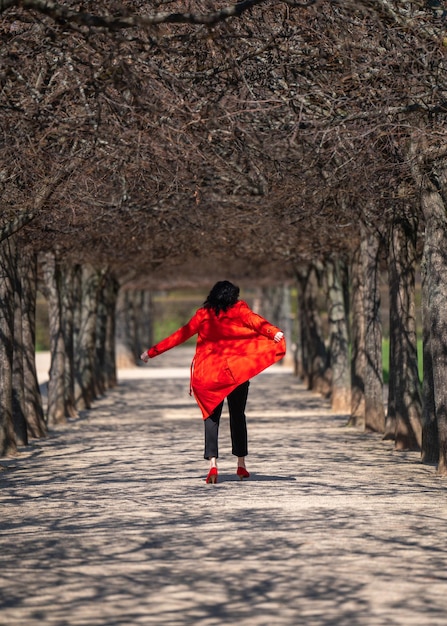 Una mujer feliz y elegante disfruta de la vida una dama de cabello oscuro con una capa roja y zapatos rojos de tacos altos camina por un callejón entre los árboles en el parque bailando vista trasera