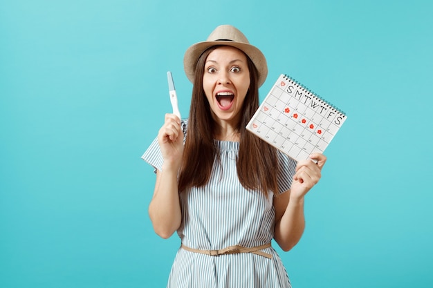 Mujer feliz emocionada en vestido azul, sombrero mantenga en la mano prueba de embarazo, calendario de períodos para comprobar los días de menstruación aislados sobre fondo azul. Concepto médico, sanitario, ginecológico. Copie el espacio.