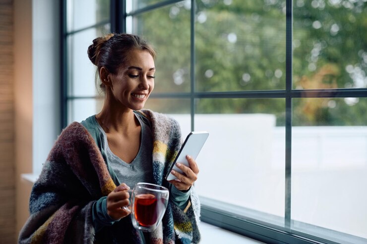 Foto mujer feliz envuelta en una manta bebiendo té mientras disfruta junto a la ventana en casa