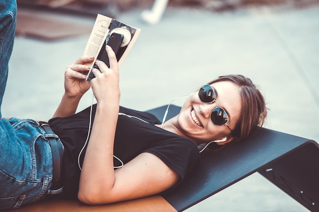 Mujer feliz escuchando música con auriculares y leyendo un libro en la calle