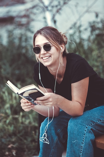 Mujer feliz escuchando música con auriculares y leyendo un libro en la calle