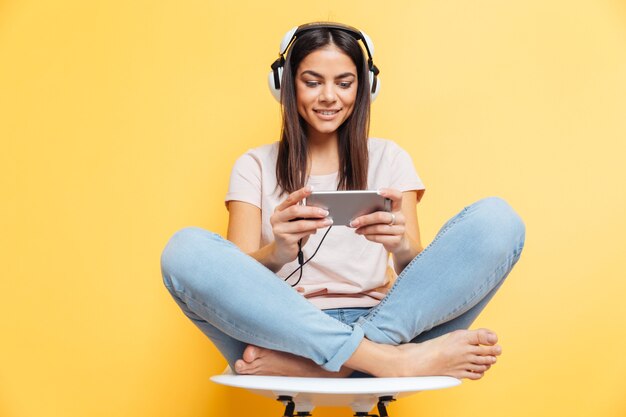 Mujer feliz escuchando música en auriculares y con smartphone sobre pared amarilla