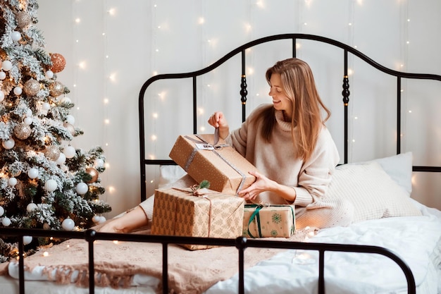 Mujer feliz está desenvolviendo cajas de regalo de Navidad o año nuevo sentado en la cama en la habitación decorada festivamente.