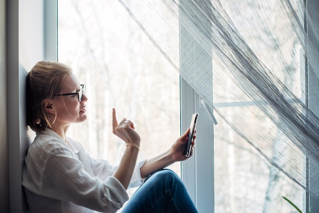 Mujer feliz con gafas y camisa blanca se sienta en el alféizar de la ventana, sosteniendo el teléfono inteligente en la mano. Linda rubia sonríe a la cámara del teléfono, hace gestos con la mano, primer plano. Chatear en línea.