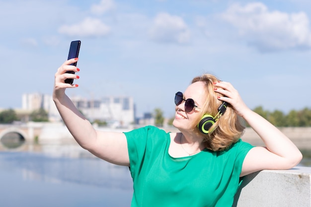 Una mujer feliz con gafas de sol y auriculares tomando una foto selfie por teléfono charlando con amigos en la calle