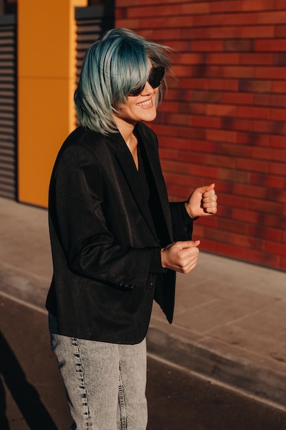 Mujer feliz con gafas de sol riendo y gesticulando mientras camina por la calle cerca del edificio de oficinas blueh